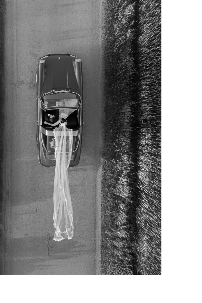 A vintage convertible carrying a bride and groom, seen from above, her veil trailing behind